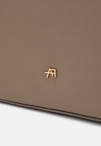 Gros plan d'une surface en cuir taupe avec un petit emblème de logo doré "A" et "F" entrelacés près du bord inférieur.