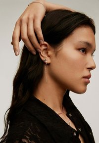 Pendientes de plata con un aro cuadrado y una cadena larga y delgada que cuelga. Combinados con una blusa de encaje oscura, resaltando su diseño elegante.