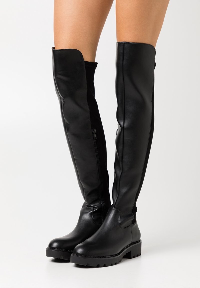Bottes en cuir noires arrivant au genou avec des fermetures éclair sur les côtés, portées sur des jambes nues, avec des semelles épaisses et des orteils arrondis sur un fond blanc.