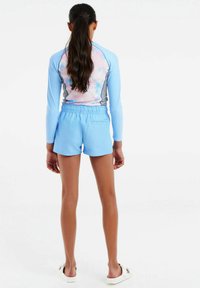 Rash guard s pastelovým tie-dye vzorom na chrbte, dlhé rukávy v svetlomodrej farbe, v kombinácii so svetlomodrými šortkami, elastickým pásom a jednoduchým dizajnom.