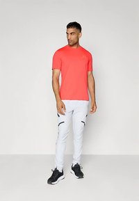 Rød atletisk t-skjorte laget av glatt materiale, kombinert med lysegrå avsmalnende bukser med svarte glidelåsdetaljer og lommer. Svarte joggesko.