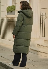 Palton pufos lung de culoare verde măsliniu, cu glugă, design matlasat și o croială lejeră. Asortat cu pantaloni negri, largi și încălțăminte casual.