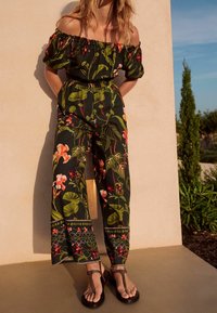 Femme portant une combinaison fleurie épaules dénudées et des sandales noires, debout contre un mur beige avec des plantes en arrière-plan.