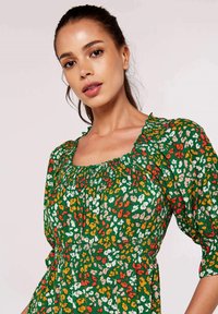 Blumenkleid mit grünem Hintergrund und orangefarbenen, weißen und gelben Mustern. Quadratischer Ausschnitt und geraffte Puffärmel. Leichter Stoff.