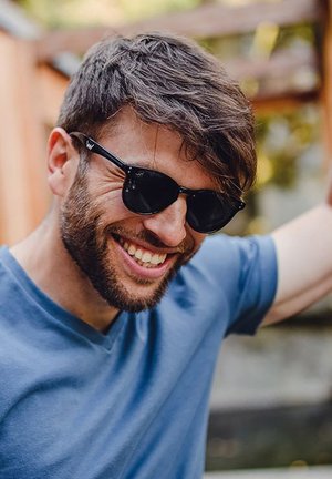 Black sunglasses with dark lenses, plastic frames, and logo detail. The person wears a blue v-neck t-shirt and has short, tousled hair.