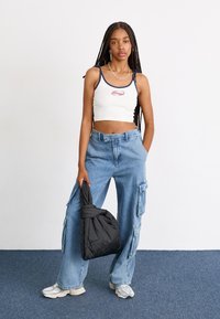 White crop top with navy trim, featuring a logo. Blue cargo jeans with side pockets. Black puffer bag. Gray sneakers. Minimalist background.