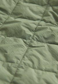 Kvilta audums bļotā zaļā krāsā ar gludu tekstūru un redzamu šuvju veidotu rombu rakstu. Viegls un mīksts izskats.