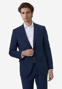 Man in een op maat gemaakt marineblauw pak met een witte shirt, die zelfverzekerd staat en met één hand zijn pakjack sluit, tegen een witte achtergrond.