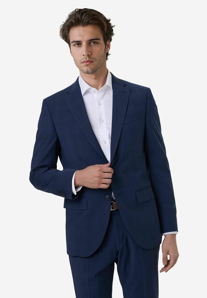 Man in een op maat gemaakt marineblauw pak met een witte shirt, die zelfverzekerd staat en met één hand zijn pakjack sluit, tegen een witte achtergrond.