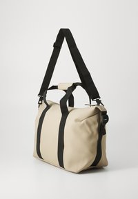 Beige duffel bag with black straps, featuring a smooth, matte texture and a structured design. Adjustable shoulder strap and dual handles.