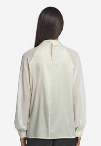 Langärmlige Bluse aus crèmefarbenem Satin mit einem Schlüssellochverschluss auf der Rückseite, gefältelten Schultern und Manschetten mit Knopf-Details. Geschmeidige, leichte Textur.