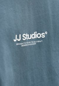 Modra bombažna majica s kratkimi rokavi z belim napisom "JJ Studios" in "Prinaša vam The J & J Crew, zasnovano na Danskem." Teksturirana tkanina.