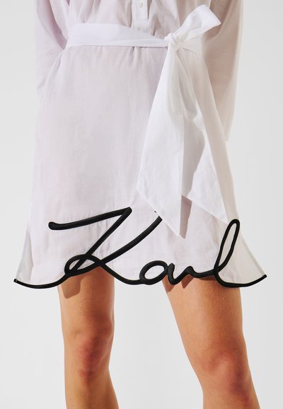 Robe en lin blanc dotée d'une ceinture à la taille et d'un détail brodé noir "Karl" le long de l'ourlet, avec une silhouette fluide.