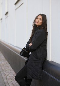 Black quilted coat with high collar, zippered side pockets, and snaps at the hem, worn with black pants and a small handbag.