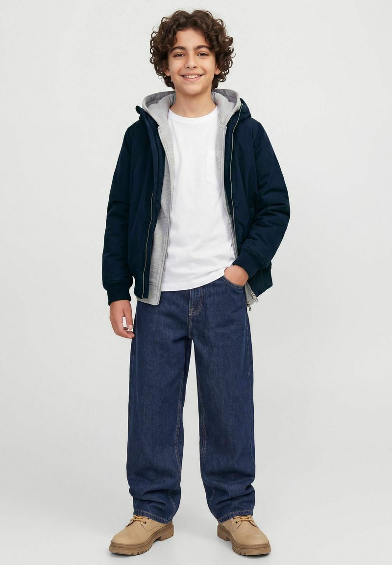 Garçon souriant aux cheveux bouclés portant une veste bleu marine par-dessus un sweat à capuche gris, une chemise blanche, un jean bleu ample et des bottes beige, debout avec une main dans la poche.