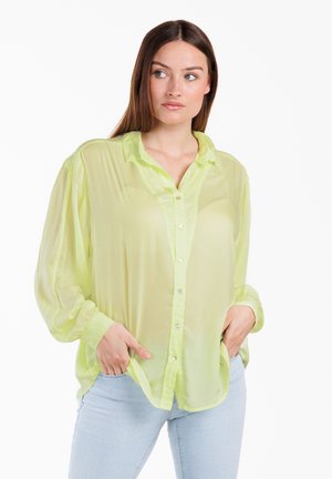 Frau mit langen braunen Haaren, die eine transparente hellgelbe Bluse mit Knöpfen und hellblaue Jeans trägt, steht mit den Händen in den Taschen und schaut zur Seite.