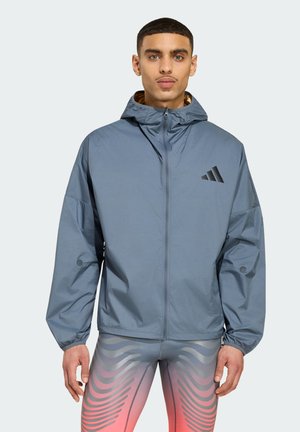 Junger Mann, der eine hellblaue Kapuzenjacke mit Reißverschluss und graue Sportleggings mit rosa und schwarzen Wellenmustern trägt, steht vor einem schlichten Hintergrund.