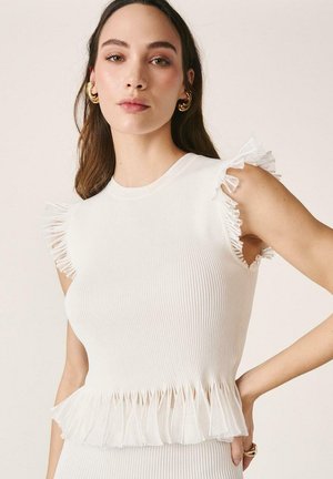 Mujer con cabello castaño que lleva pendientes de aro dorados y una camiseta blanca sin mangas con textura acanalada y bordes con volantes en los hombros y la cintura.