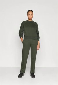 Sieviete stāv ar rokām kabatās, tērpta tumši zaļā sporta džemperī, atbilstošās biksēs un melnās sporta apavās, pret vienkāršu gaismu fona.