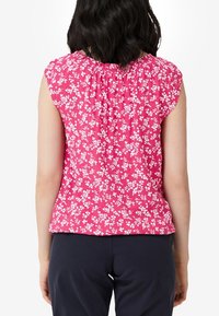Blusa de manga corta, rosa con estampado floral y patrones florales blancos, con cuello acanalado y corte holgado. Llevada con pantalones de color oscuro.