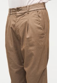 Hnědé kalhoty s plisováním, vedlejšími kapsami a pasem na knoflíky, nošené s vsunutou světle růžovou košilí.