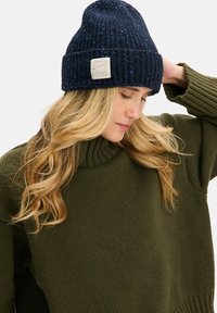 Granatowa dzianinowa czapka z prążkowaną teksturą, z metką. Połączona z grubym oliwkowozielonym swetrem z wysokim kołnierzem i luźnym krojem.