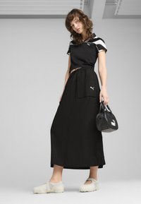 Juodas trumpas marškinėliai su baltais akcentais, aukštu liemeniu juoda sijonas su kišenėmis, pilki slip-on tipo bateliai su tekstūruota sole, juoda rankinė su logotipu.