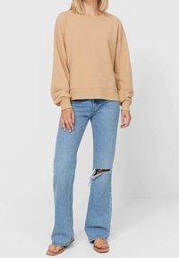 Sweatshirt beige surdimensionné avec des manches longues, associé à un jean évasé bleu clair avec un déchirure au genou, et des sandales tan simples.
