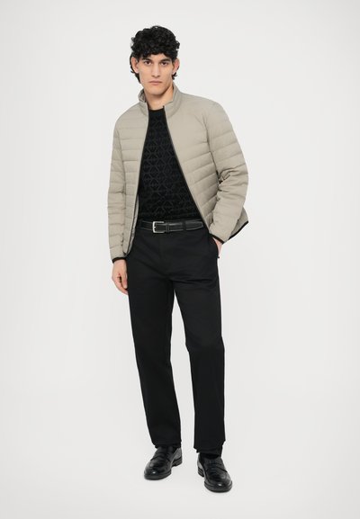 Svetlo siva prešita jakna čez črno srajco s potiskom, kombinirana s črnimi hlačami in sijočimi črnimi loaferji. Model stoji z rokami v žepih.