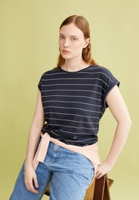 Marine gestreepte t-shirt met opgerolde mouwen, gecombineerd met lichtblauwe spijkerbroek en een zachtroze geribbelde riem geknoopt om de taille. Lichtgroene achtergrond.