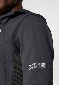 Black athletic jacket with a fitted design, featuring textured mesh paneling and Eiger Extreme logo on the left sleeve in white.