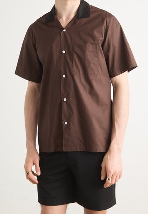 Mann trägt ein braunes Kurzarmhemd mit schwarzen Kragen und schwarze Shorts, steht vor einem einfarbigen Hintergrund.