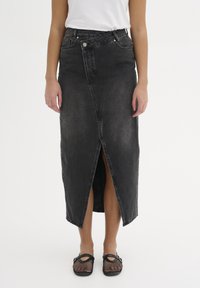 Gonna midi in denim nera con uno spacco frontale, presenta una vita con passanti per cintura e tasche anteriori. Indossata con scarpe in mesh nere.