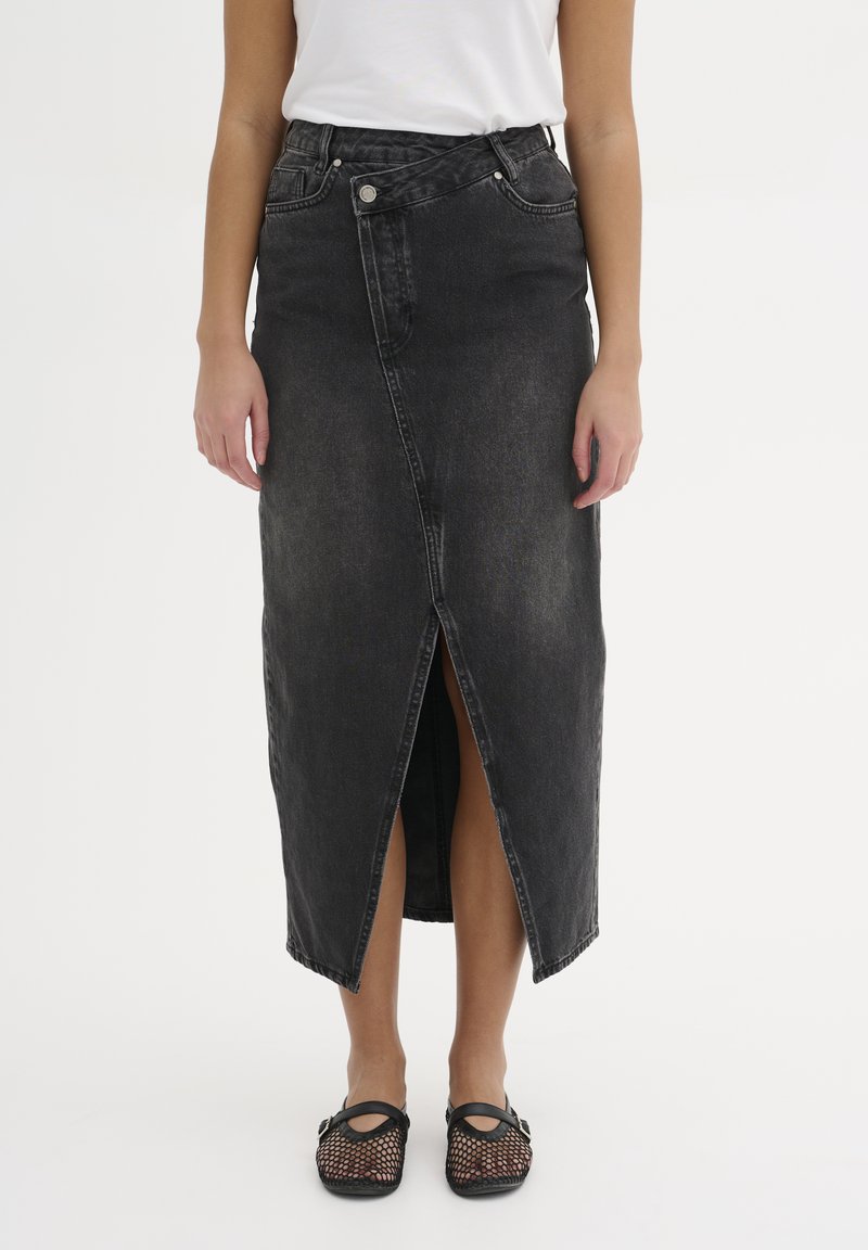 Gonna midi in denim nera con uno spacco frontale, presenta una vita con passanti per cintura e tasche anteriori. Indossata con scarpe in mesh nere.