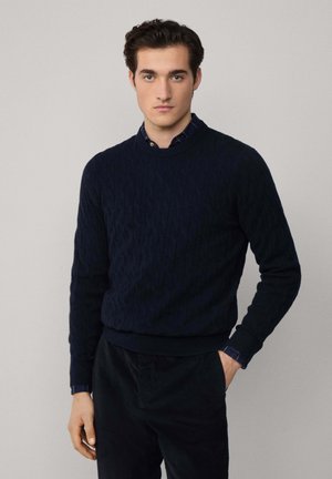 Un pull en jersey de couleur bleu marine avec un col rond et des poignets et un ourlet côtelés, associé à un pantalon sombre et une chemise à col en dessous.
