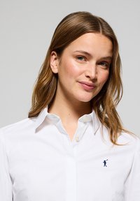 Camicia bianca con bottoni in tessuto liscio, dotata di colletto classico. Presenta un logo blu di un golfista ricamato sul lato sinistro del petto.