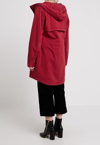 Giacca rossa con cappuccio, dal taglio rilassato e dettagli sulla schiena, abbinata a pantaloni neri a vita alta e stivaletti neri con tacco.