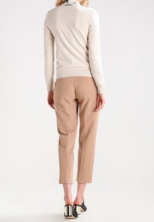 Femme portant un pantalon beige court, un pull crème et des talons noirs à talons blocs métalliques, debout de dos sur un fond blanc.