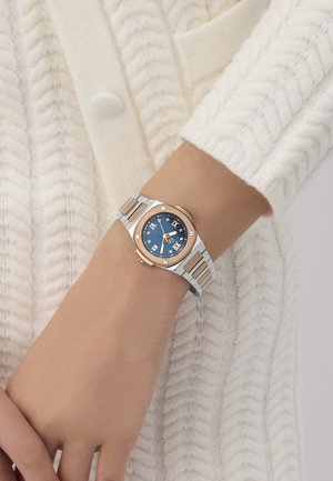 Sudraba un rozā zelta pulkstenis ar zilu ciparnīcu, romiešu cipariem un metāla saišu aproci, uz rokas, kas atrodas pretī teksturētai krēmsvītrai džempera.