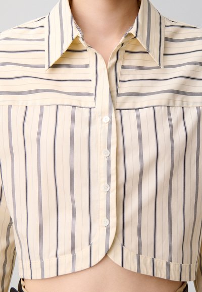 Camicia corta con bottoni in crema chiaro con righe verticali blu navy, con colletto e piccoli bottoni lungo la patta anteriore.