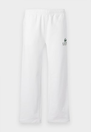 Deux personnes se tiennent côte à côte, portant des chemises vertes Lacoste et des pantalons blancs Lacoste, sur un fond blanc uni.
