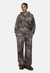 Sweat à capuche camouflé raccourci et pantalon ample dans des tons de marron et de gris avec des motifs de feuilles. Comprend des poches et une capuche ajustable.