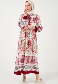Largo vestido floral en blanco con patrones rojos y verdes. Cuenta con mangas gathered y un dobladillo con acentos decorativos. Se lleva con zapatillas beige.