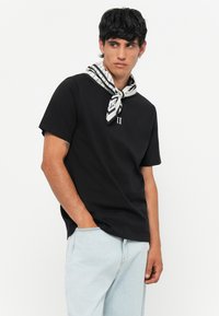 Camiseta de algodón negra de manga corta, con un cuello redondo. Una bufanda a rayas enrollada alrededor del cuello añade contraste y detalle. Jeans de mezclilla clara.