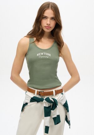 Débardeur côtelé vert olive avec le texte blanc "New York", associé à un pantalon crème et un sweat-shirt rayé vert noué à la taille. Ceinture marron.