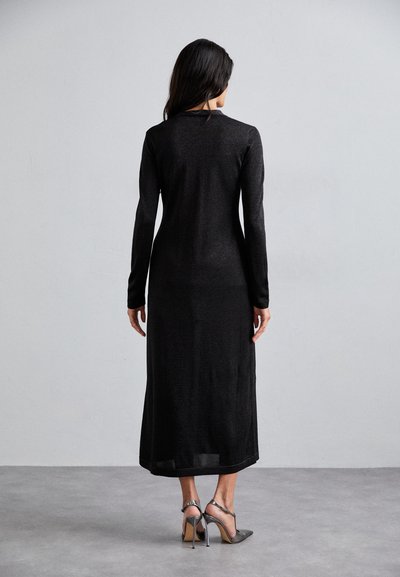 Vestido negro de mangas largas en un material de punto elegante, con una silueta recta y longitud a mitad de pantorrilla. Combinado con zapatos de tacón alto plateados.