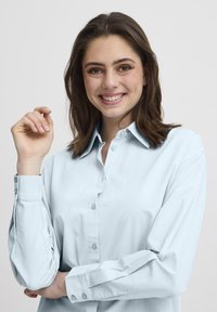 Camisa azul claro de botones con cuello, que presenta una textura suave y mangas largas, con un brillo sutil. Detalles de botones en los puños.