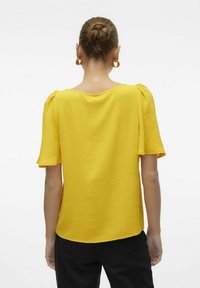 Kollane blouse, millel on tekstuuriga kangas, lühikesed puffvarrukad ja laia kaelus. Kandmine mustade pükste juurde, mis näitavad mugavat istuvust.