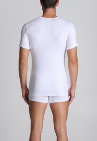 Uomo in piedi di spalle che indossa una maglietta bianca attillata a maniche corte e slip bianchi coordinati, su uno sfondo grigio uniforme.