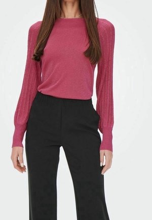 Femme portant un haut rose cintré à manches longues bouffantes et un pantalon noir taille haute à jambes larges, posant devant un fond uni.